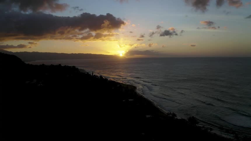 The sun peaks over distant mountains to shine on the long coast of a landmass at sunrise