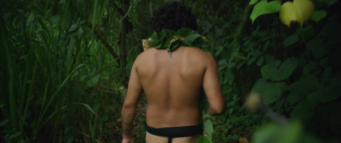 A man with a loincloth and leaves around his shoulders walking in a forest with plants and trees