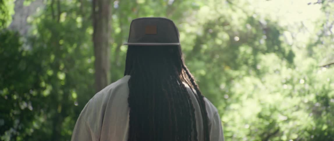 A man with dreadlocks and a hat walks through the forest in midday