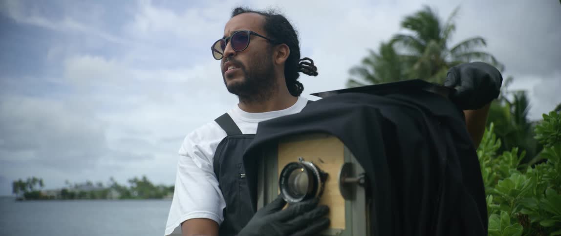 Bearded man adjusts a zoom lens on a vintage camera while standing next to a river