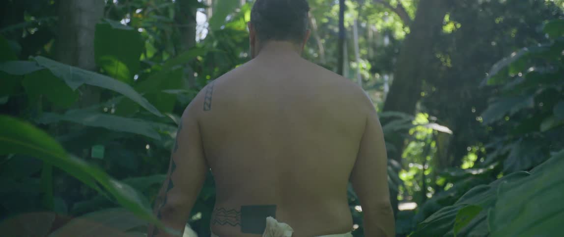A bare chested man walks through the jungle with a white cloth around his waist