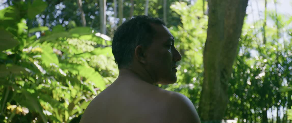 A bare chested man stands in nature looking around during the day