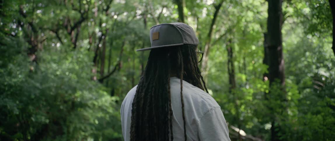 A black man with dreadlocks hat and glasses stands inside a forest during the day