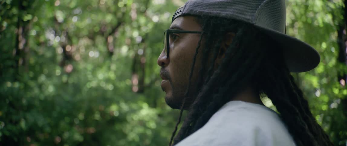 A black man with dreadlocks and glasses stands in a forest