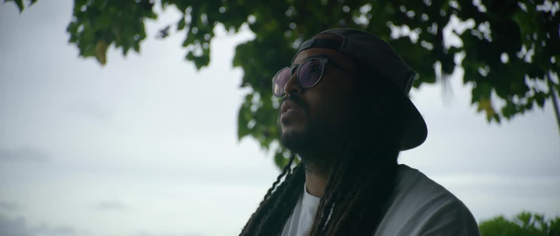 A man with sunglasses and a hat with trees the sky and clouds in the background