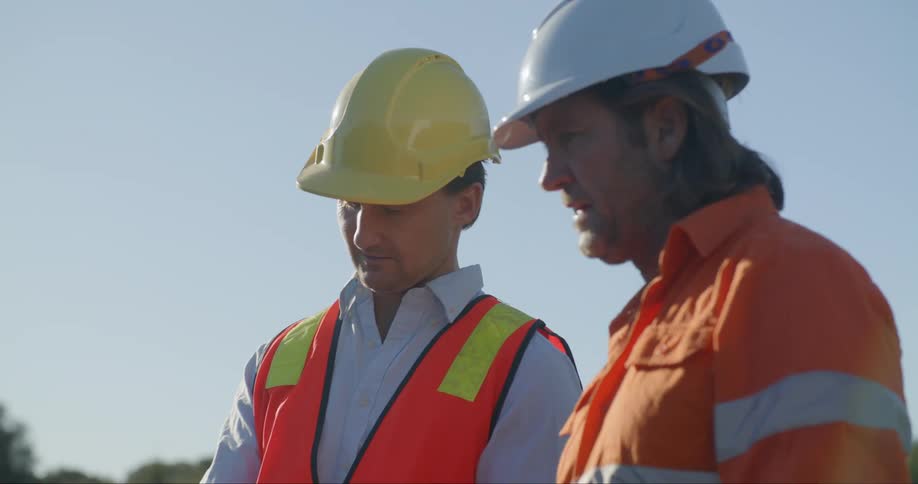 A pair of men in reflective safety clothing and hard hats speak with one another while standing outdoors on a sunny day