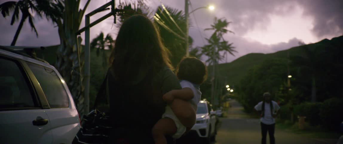 A woman carries a baby with one arm as she walks across a road in a neighborhood towards a waiting man and child
