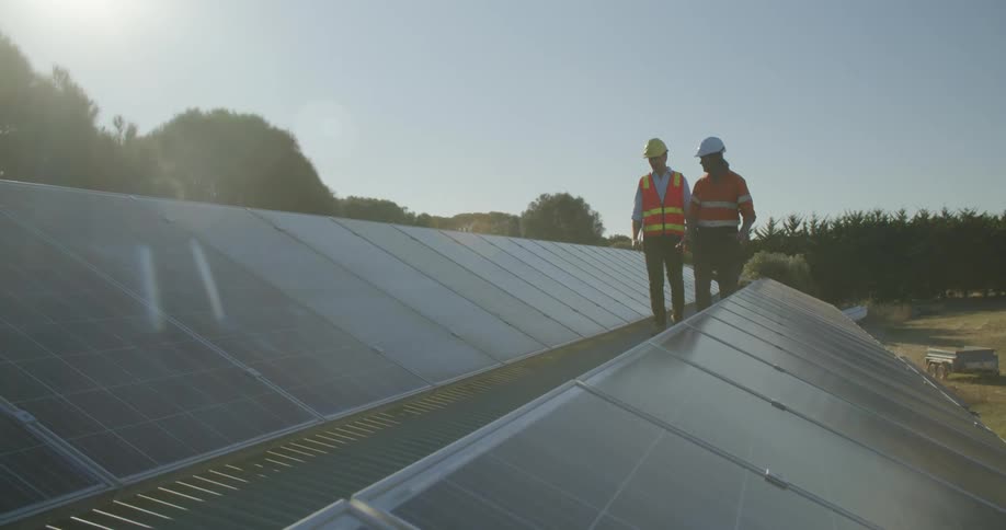Two men wearing construction gear walking on a platform between two rows of solar panels up off the ground