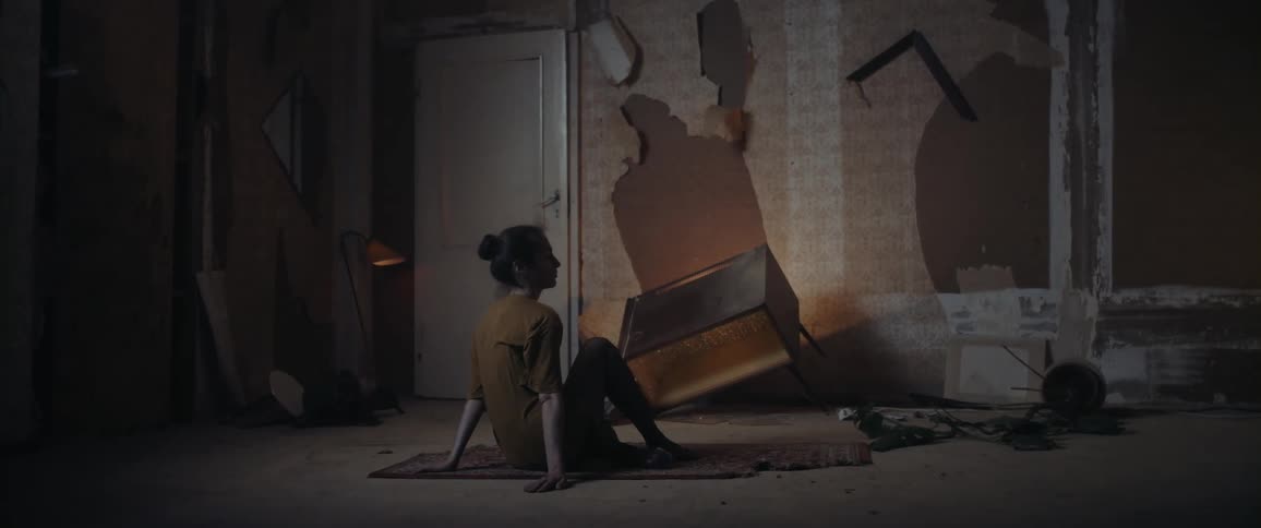 A young woman runs her hand across the floor as she sits on a worn rug in a dilapidated room with flickering lamps