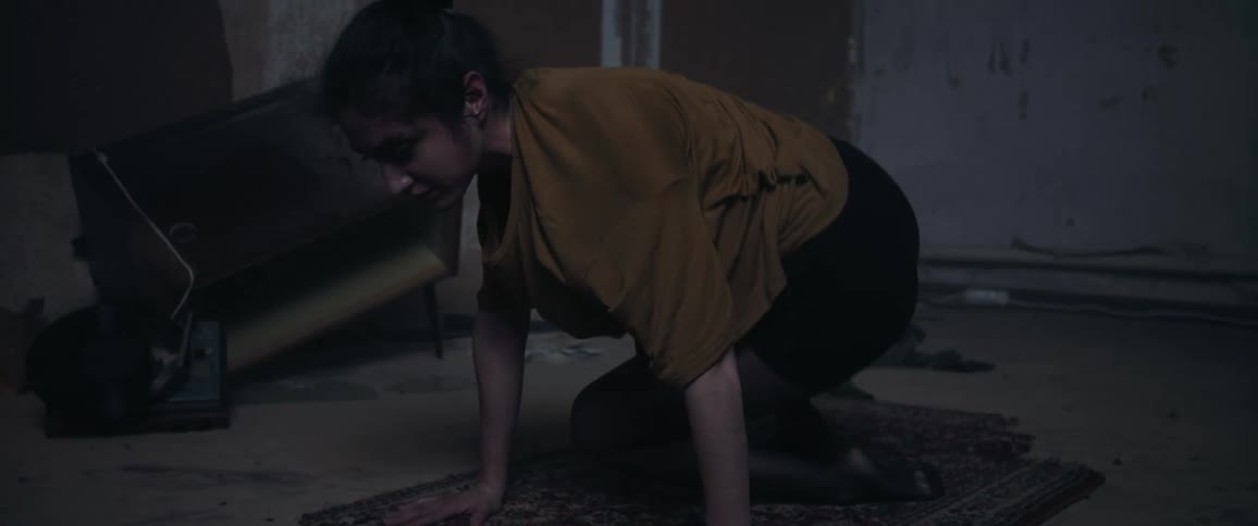 Light flashes in an abandoned room while a woman performs a dance pose on a rug and lowers herself to the floor