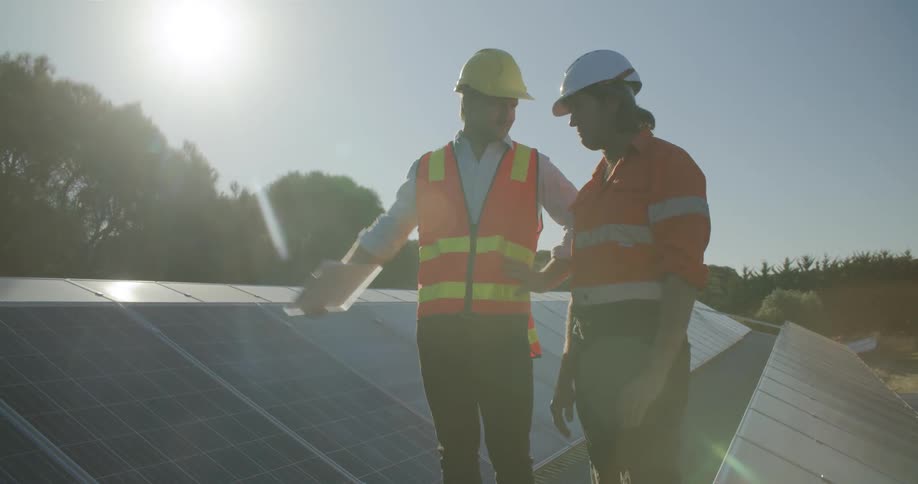 Two construction workers on a rooftop with solar panels having a discussion on a sunny day