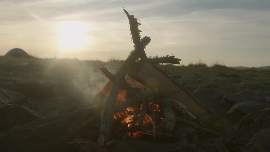 Campfire burns and flames flickering as the sun sets in a cloudy sky