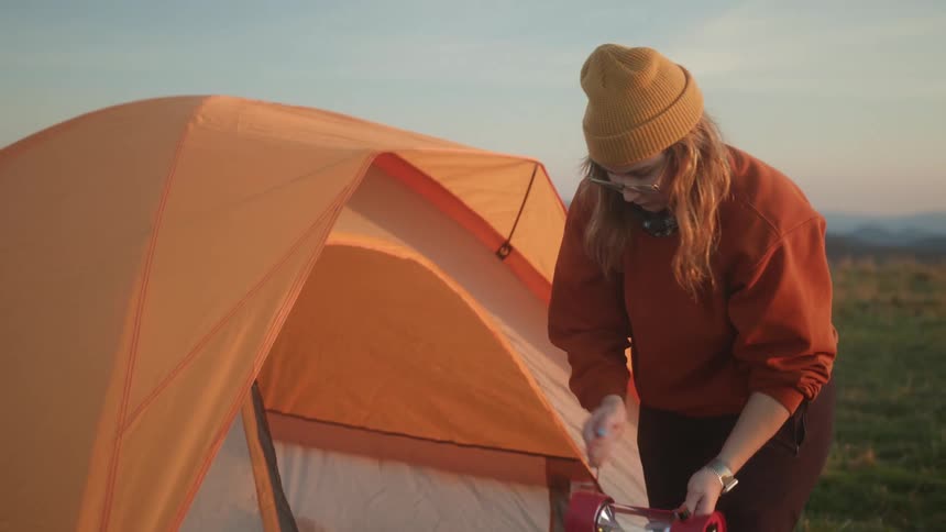 A woman with a hat and glasses looking at a lantern in front of a tent with the sky and clouds in the background