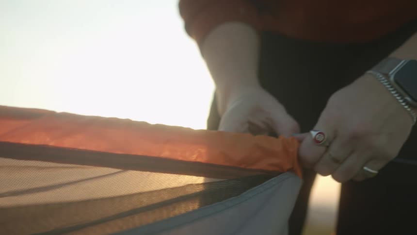 A woman threads a pole into an orange tent in a field