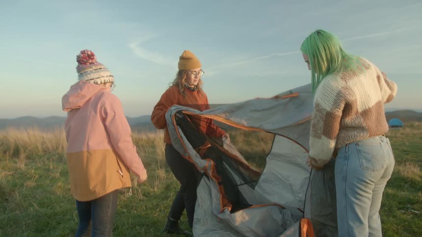 Three young women fold out a tent for camping in a large field near a mountain