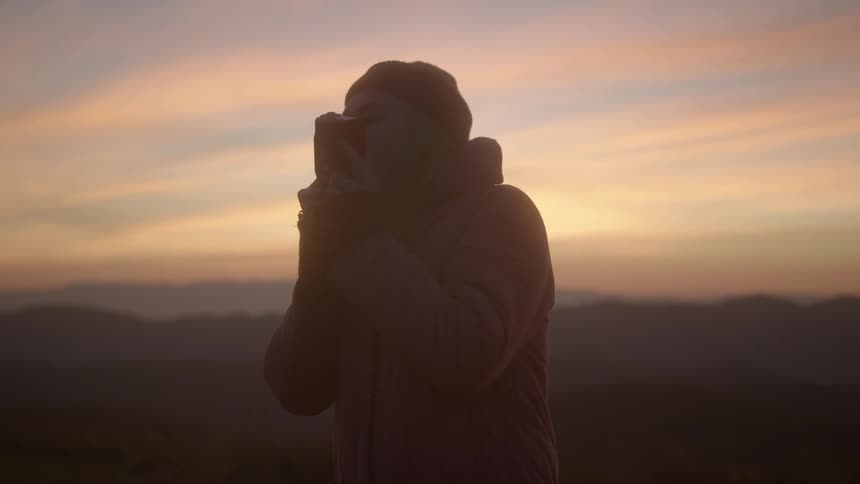 A female photographer in a coat taking a photograph outside at sunset