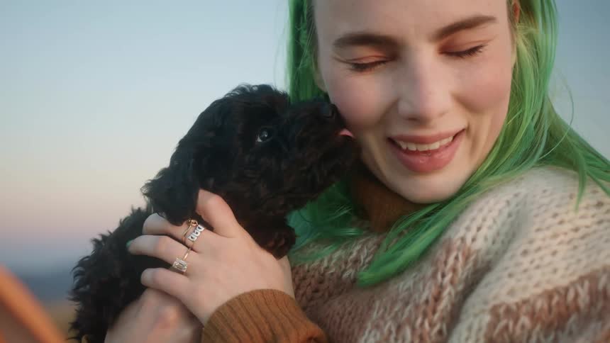 A green haired female holding a black puppy that is licking her face