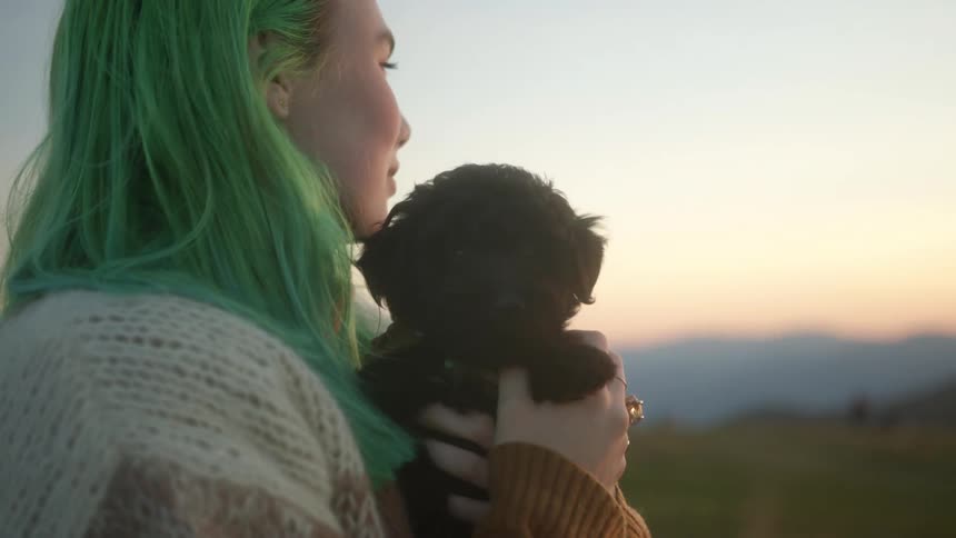 A woman with green hair holding and kissing a puppy with a landscape sky and clouds in the background