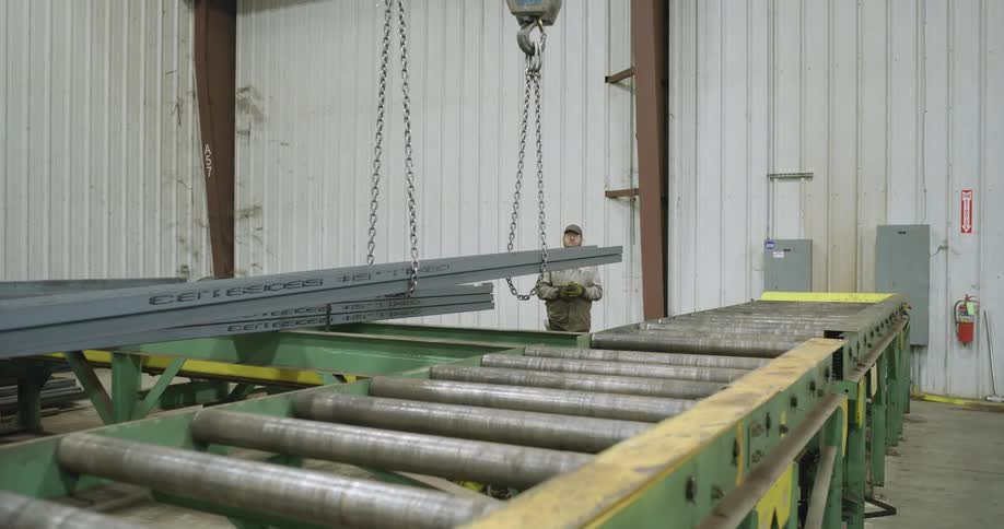 A man at a factory watches as steel is lowered to a conveyor belt