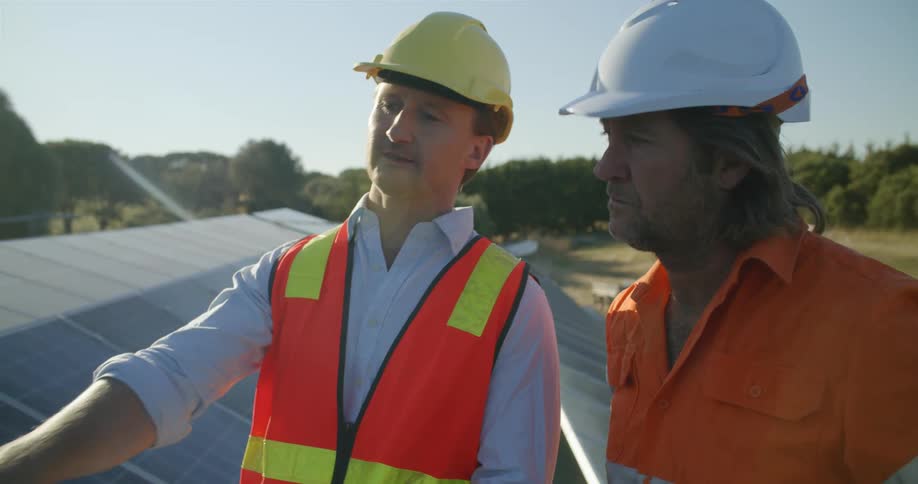 Two men discuss a project outdoors wearing hard hats and a safety vest