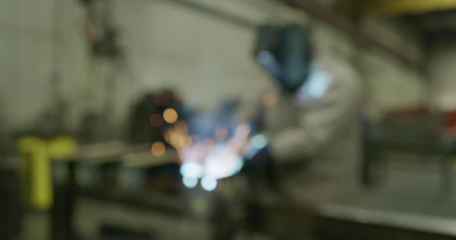 Out of focus shot of a worker soldering metal profiles over a bench in a factory inside a warehouse