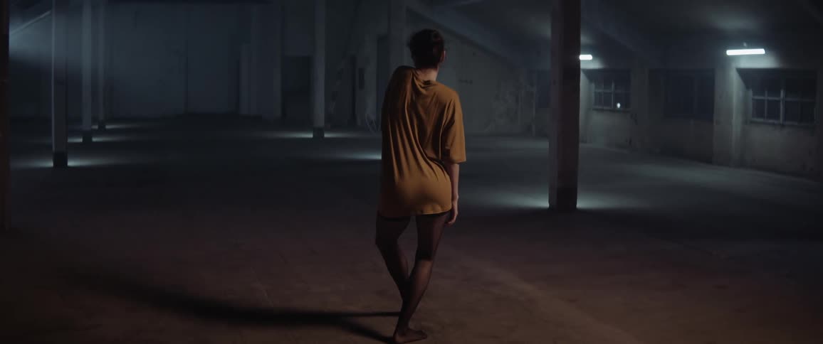 A woman makes slow graceful dancing movements in an empty dimly lit building