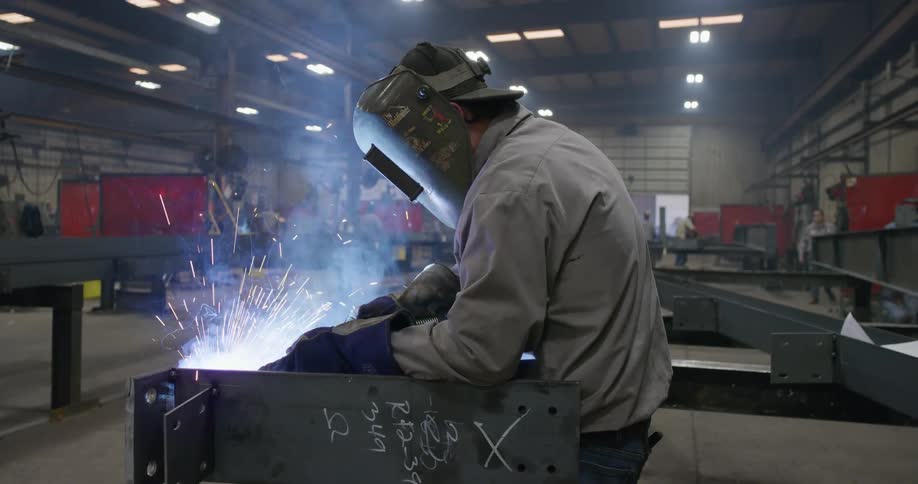 A man welding in a factory as his boss and coworker walk in the back ground