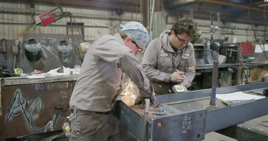 A man takes measurements as another man uses a tool to weld a piece of metal into place inside of a warehouse