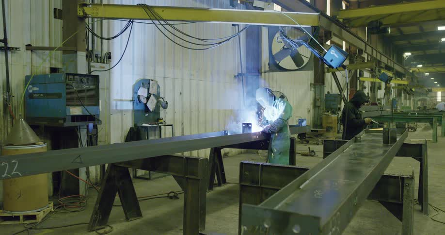 Workers wearing soldering mask solders steel profiles over a bench in a factory inside a warehouse