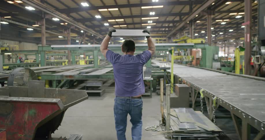 A man working in a warehouse grabs a slab of metal from next to a man holding a clipboard and walks it over to a structure and throws it
