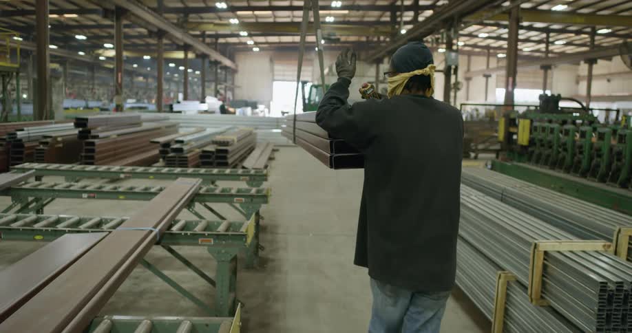 A warehouse employee walks and guides a set of steel beams that are tied together from above
