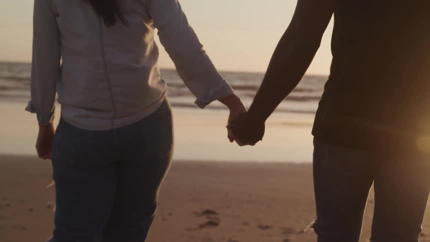 A man and a woman walking on the beach and holding hands with the ocean sky and clouds in the background