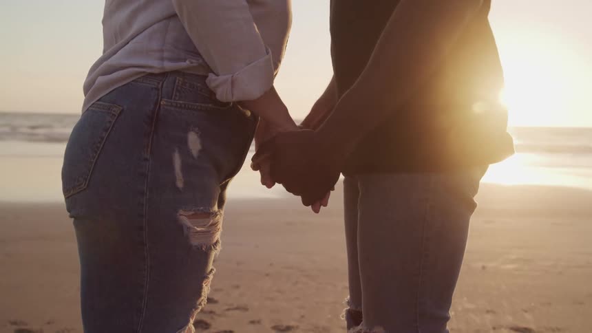 A man and a woman standing in a beach holding hands with the ocean the sky and clouds in the background