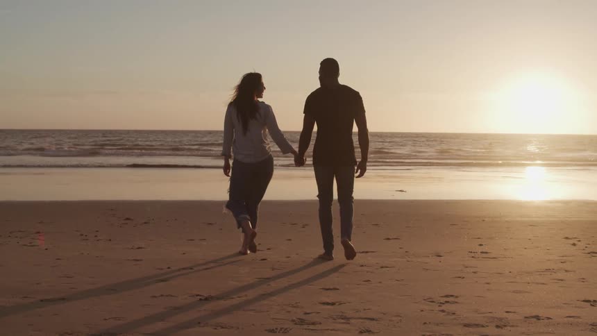 Couple holding hands walk over the sand of a beach next to the ocean at sunset