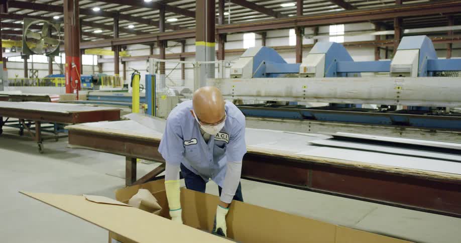 A factory worker places a long piece of white sheet metal in a cardboard box