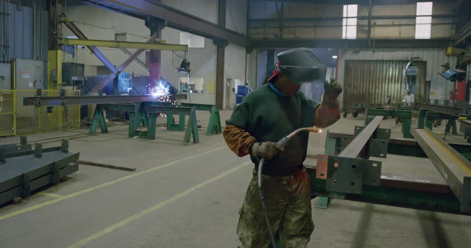 A man in personal protective equipment uses a welding torch on a metal beam in a factory during the day