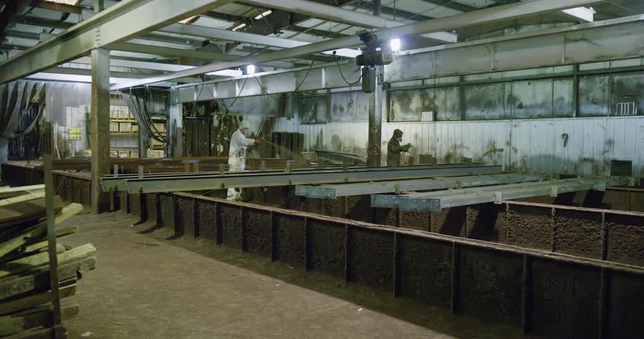 Workers wearing protective gear spray metal beam structures in factory