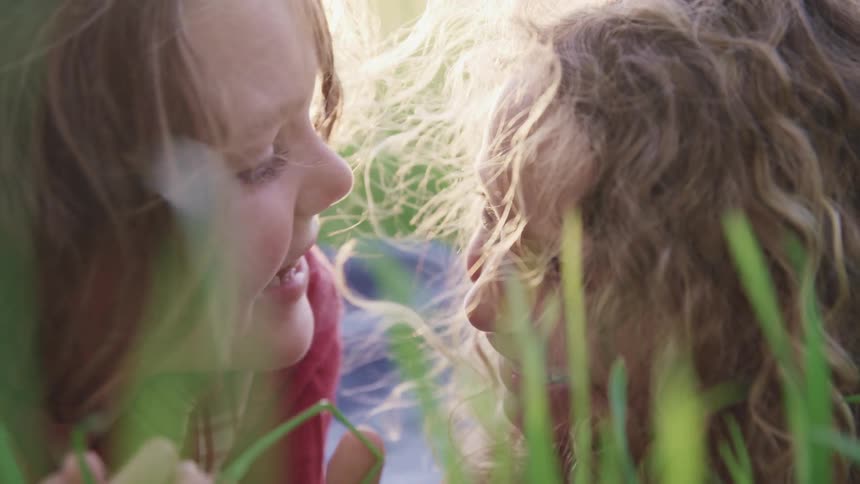 A boy and girl lying side by side on their stomachs in a field of grass smile into the camera before turning to smile at one another
