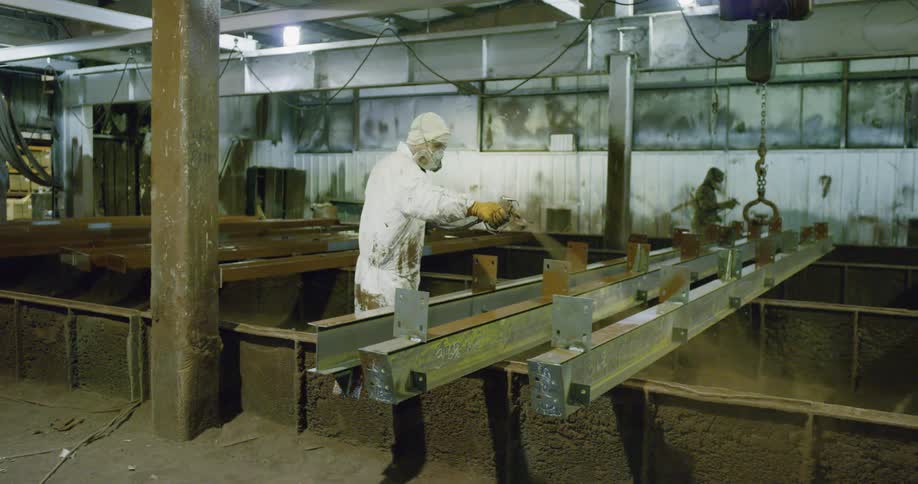 Factory workers wearing protective gear use a heavy duty spray to clean steel beams