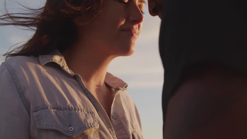 A young woman stands outside with her eyes closes as a man moves in to kiss her