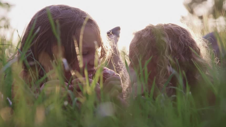 A boy and girl lying side by side in a field during the day turn to look at each other before beginning to laugh