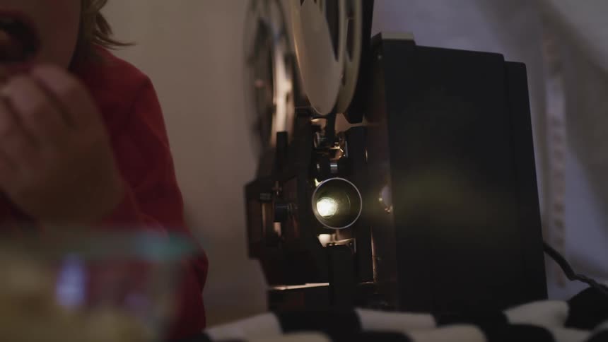 A woman lies down next to a movie projector and eats popcorn while watching a film at home