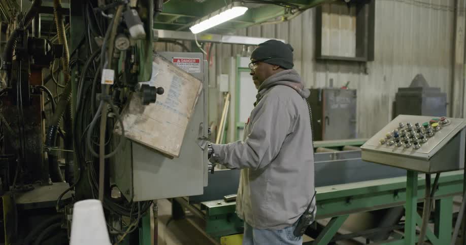 A man with a hat and gloves and a walkie talkie working in a factory next to a conveyor belt