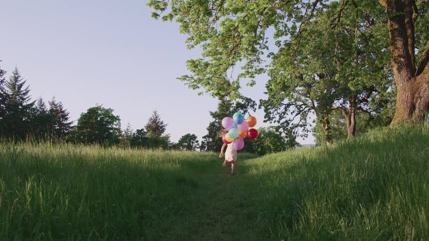 Young girl wearing a dress is running through the grass with a bunch of colored balloons and trees all around