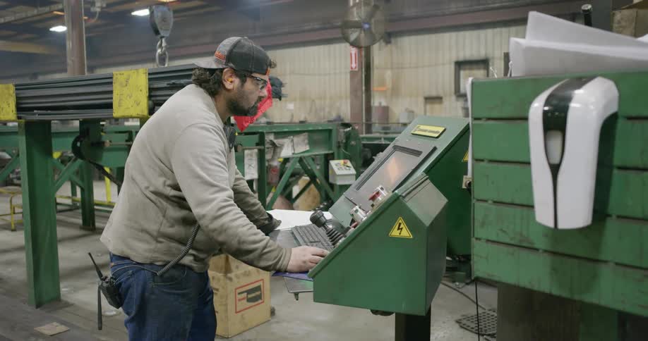 Man works on a screen in front of a machine inside of a warehouse then picks up a glove and puts it on
