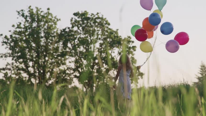 Young girl wearing white dress carries a bunch of colored balloons as she walks through the grass towards trees