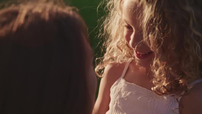 A young girl with curly blonde hair smiles widely at her mother as they stand outside