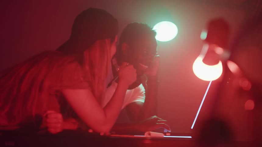 A woman leans on her coworkers desk in a dimly lit studio as they look at a computer screen together