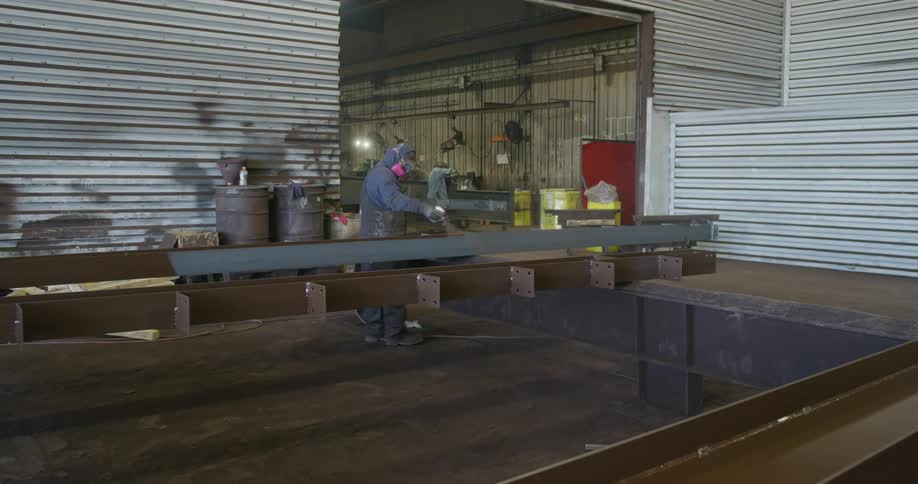 Two men working inside of a factory warehouse building on long pieces of steel wearing safety suits and masks