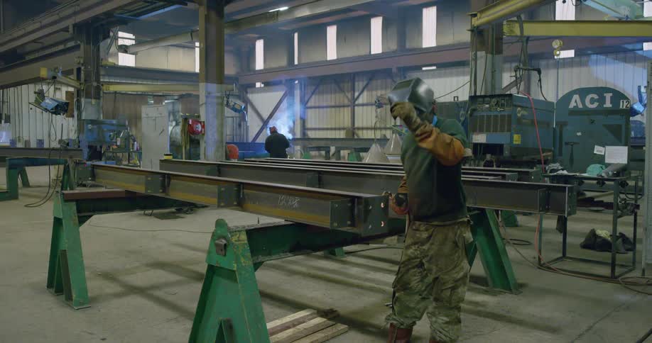 A man in personal protective equipment uses a welding torch on a metal beam in a factory during the day